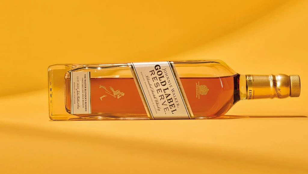 Garrafa de whisky Johnnie Walker Gold Label deitada em um fundo amarelo.