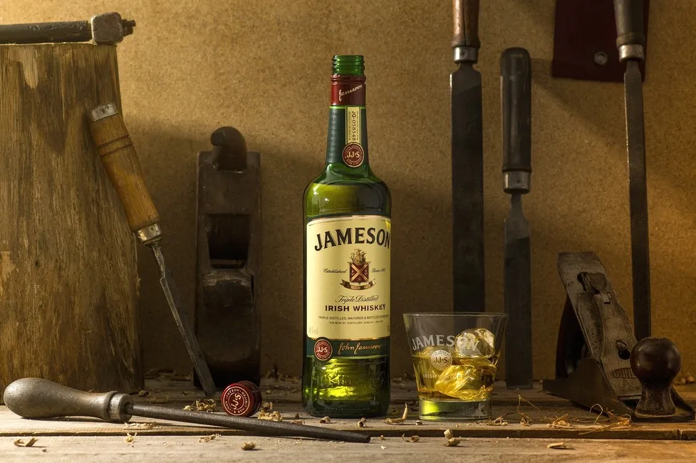 Garrafa de whisky Jameson ao lado de um copo com whisky e gelo, com ferramentas de carpintaria ao fundo