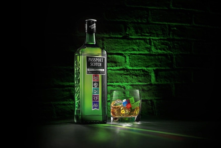 Garrafa de whisky Passport ao lado de um copo com whisky e gelo, com uma parede verde iluminada ao fundo