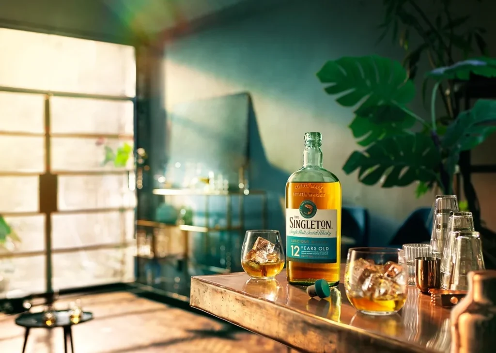 Garrafa de whisky Singleton 12 anos ao lado de dois copos com whisky e gelo, em um ambiente iluminado com plantas ao fundo.