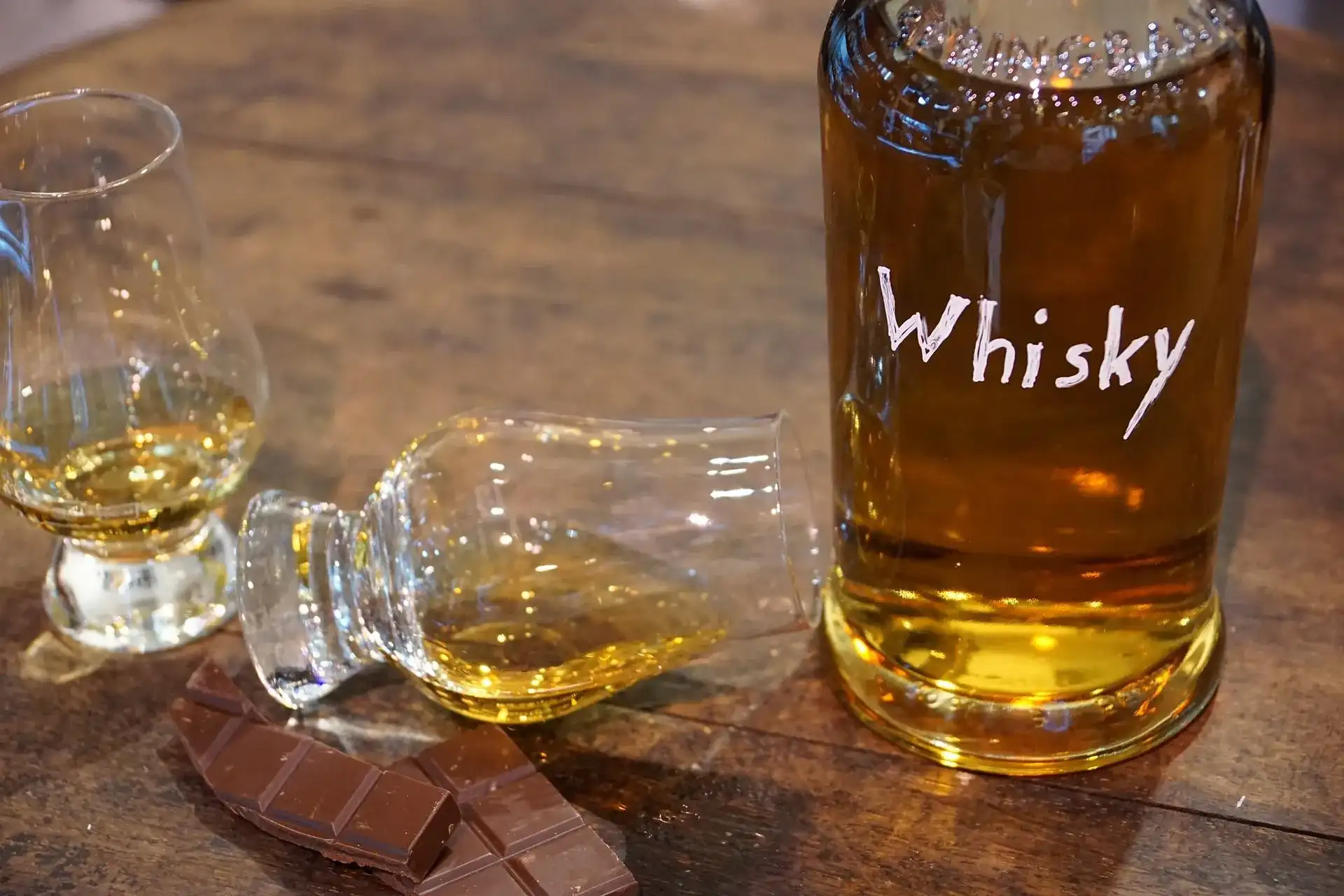 Garrafa de whisky com um copo Glencairn tombado e uma barra de chocolate, ilustrando os melhores whiskys até 500 reais.