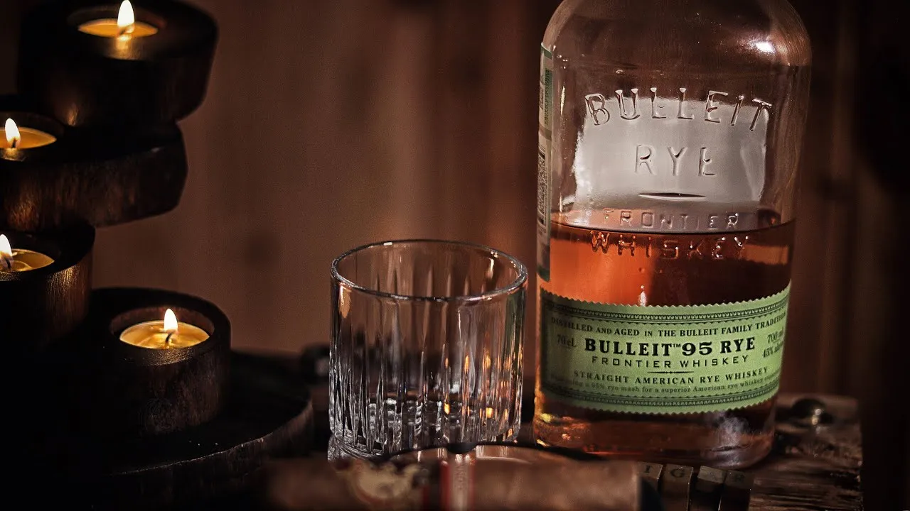 Garrafa de Bulleit Rye ao lado de um copo vazio sobre uma mesa, destacando o whisky de centeio.