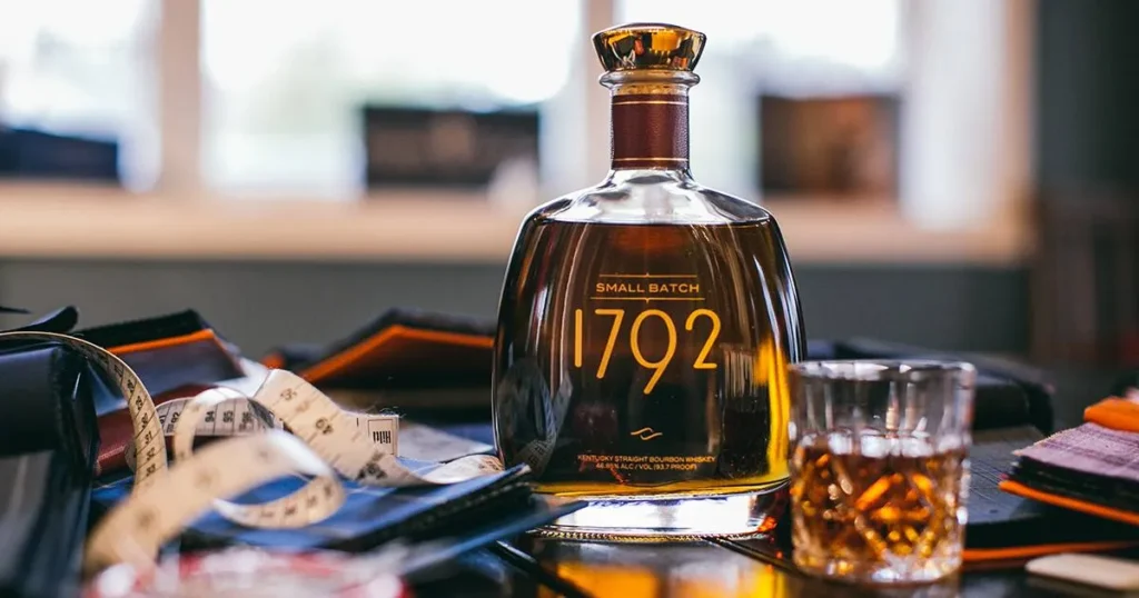 Garrafa de Whisky 1792 Small Batch ao lado de um copo com bourbon, em um ambiente sofisticado com tecidos e fita métrica.
