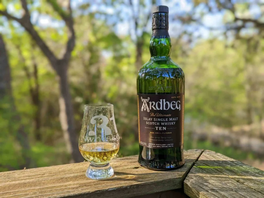 Garrafa de Whisky Ardbeg 10 Anos e um copo com a bebida sobre uma superfície de madeira, com um cenário natural ao fundo.