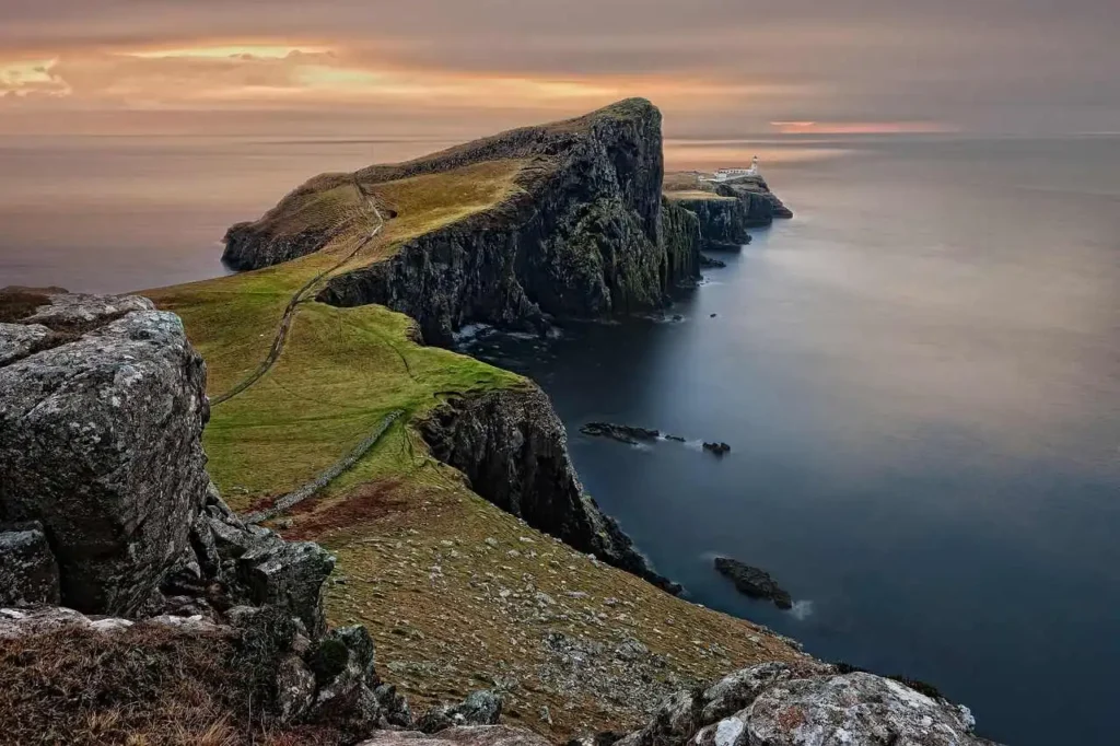 Paisagem costeira rochosa da Escócia próxima à destilaria do Whisky Talisker 10 Anos, com penhascos e farol ao fundo